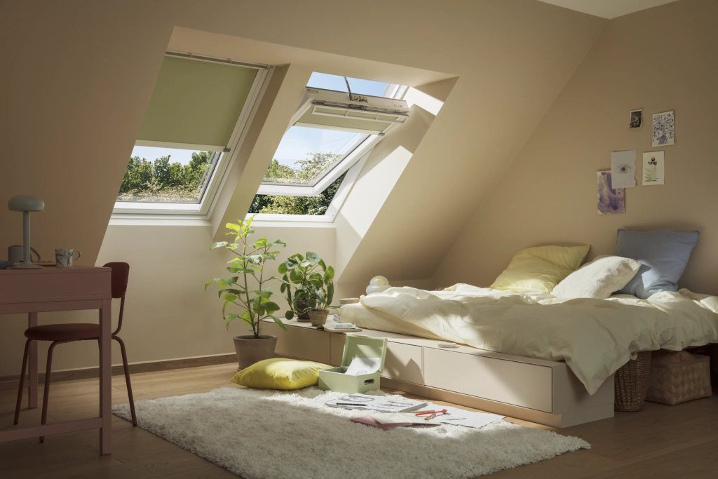 Cozy attic bedroom with skylight windows, low platform bed with cream bedding, potted plants, desk, and shaggy white rug.