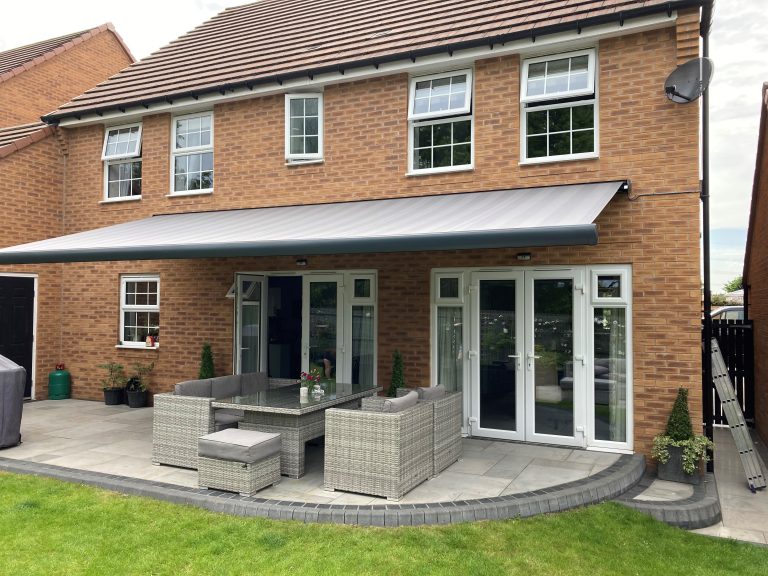 Rear view of brick house with retractable awning over patio, rattan furniture set, and French doors leading to garden.