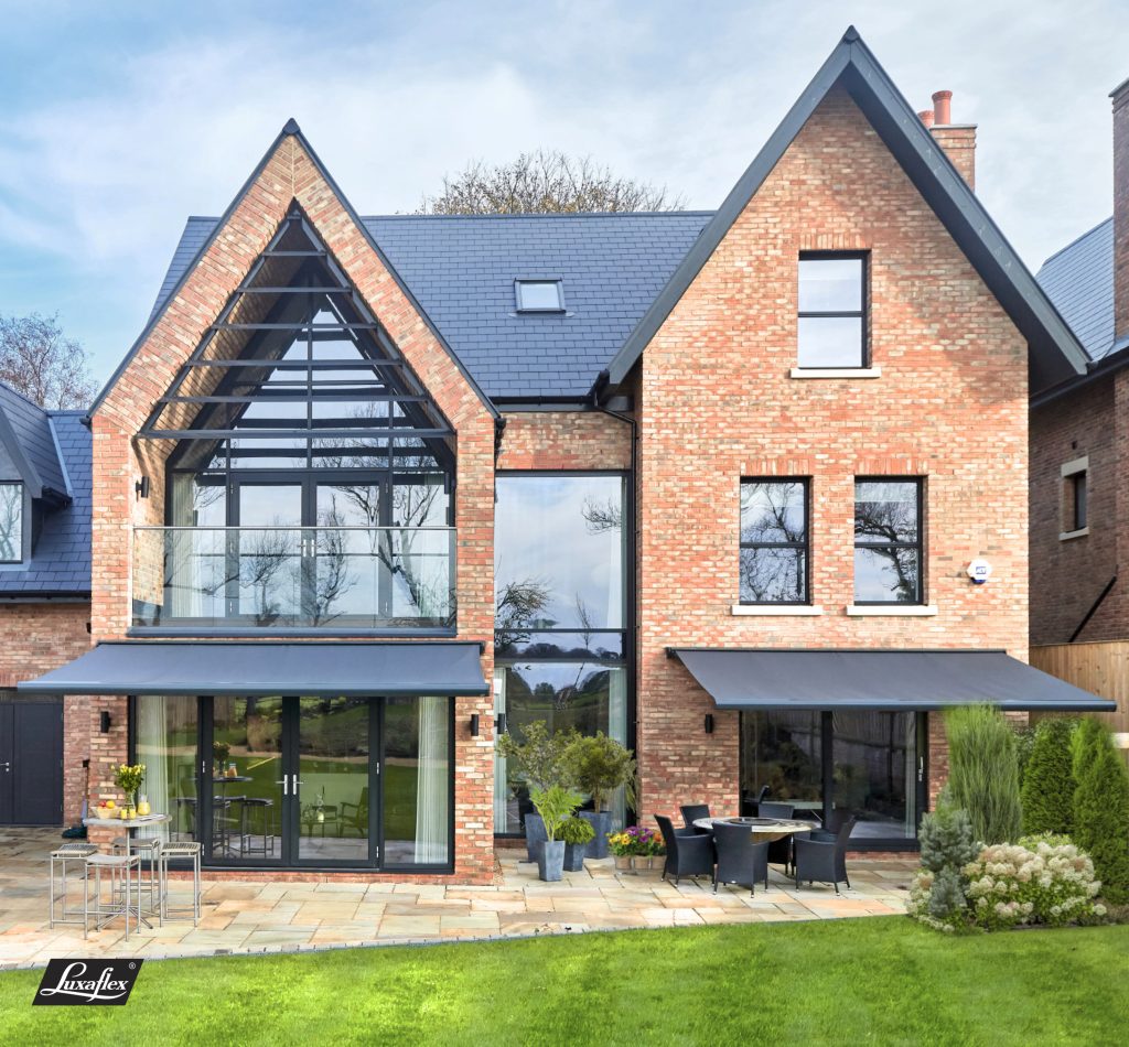 Modern brick house with dual gables, large glass windows, retractable awnings, patio furniture, and landscaped garden.