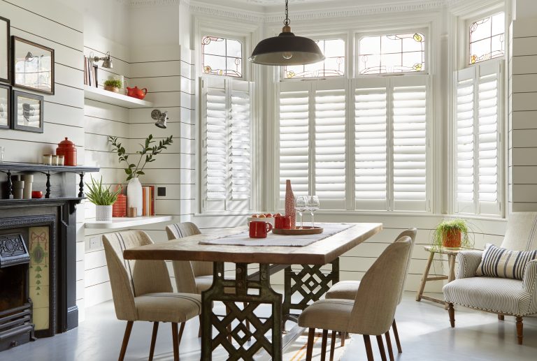 Bay window dining room with white plantation shutters, stained glass, wood table, and vintage cast iron fireplace.
