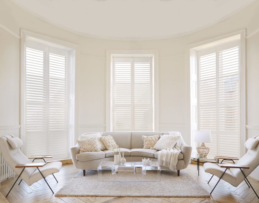 Elegant living room with white plantation shutters, curved sofa, mid-century chairs, and acrylic coffee table on herringbone floor.