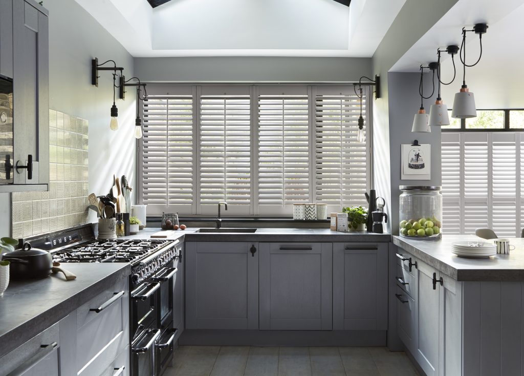 Gray U-shaped kitchen with plantation shutters, Smeg range cooker, industrial pendant lights, and stone countertops.