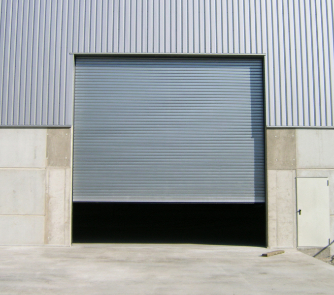 Industrial warehouse with metal roller shutter door partially open, concrete base panels, and small side entry door.
