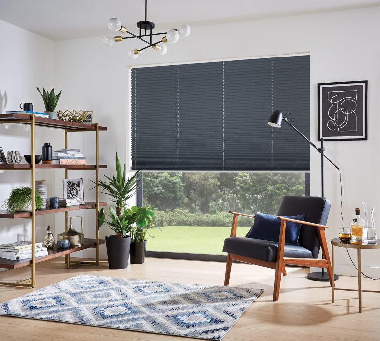 Modern living room with navy blue accent chair, gold-framed bookshelf, patterned rug, and pleated shade window covering.