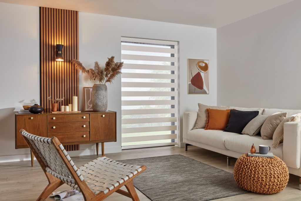 Modern living room with beige sofa, wood slat wall, zebra blinds, woven lounge chair, and knit orange pouf.