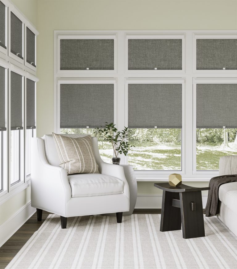 White wingback chair with striped pillow beside grey roller shades, wood side table, and dark sofa in bright sunroom.