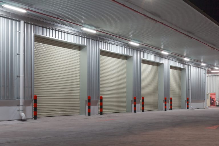 Warehouse loading dock at night with closed roller shutter doors and red-black safety bollards