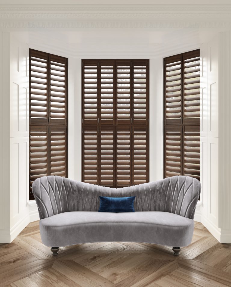 Curved gray velvet sofa with navy pillow in bay window nook with dark wood plantation shutters and herringbone floor.