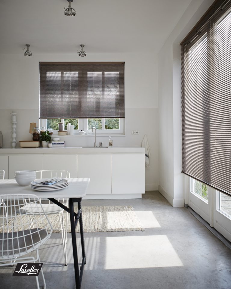 Modern white kitchen with venetian blinds on windows, wire chairs, minimalist dining table, and concrete flooring.
