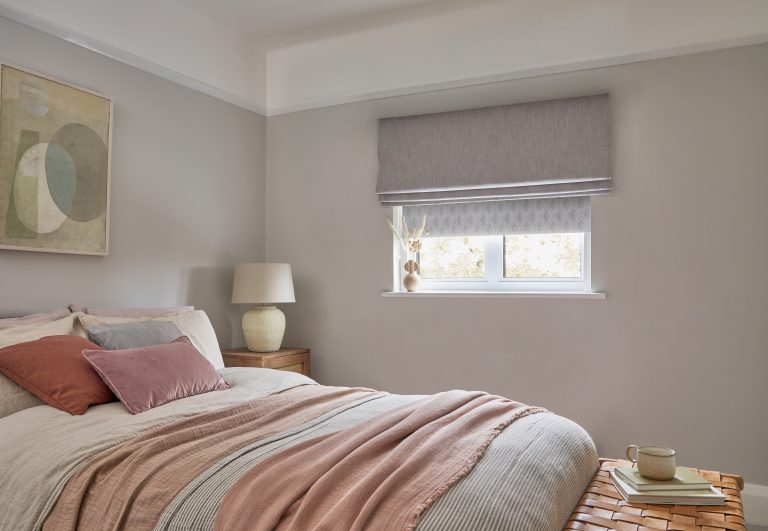 Cozy bedroom with gray walls, pink and beige bedding, layered Roman shades, abstract art, and warm wooden accents.
