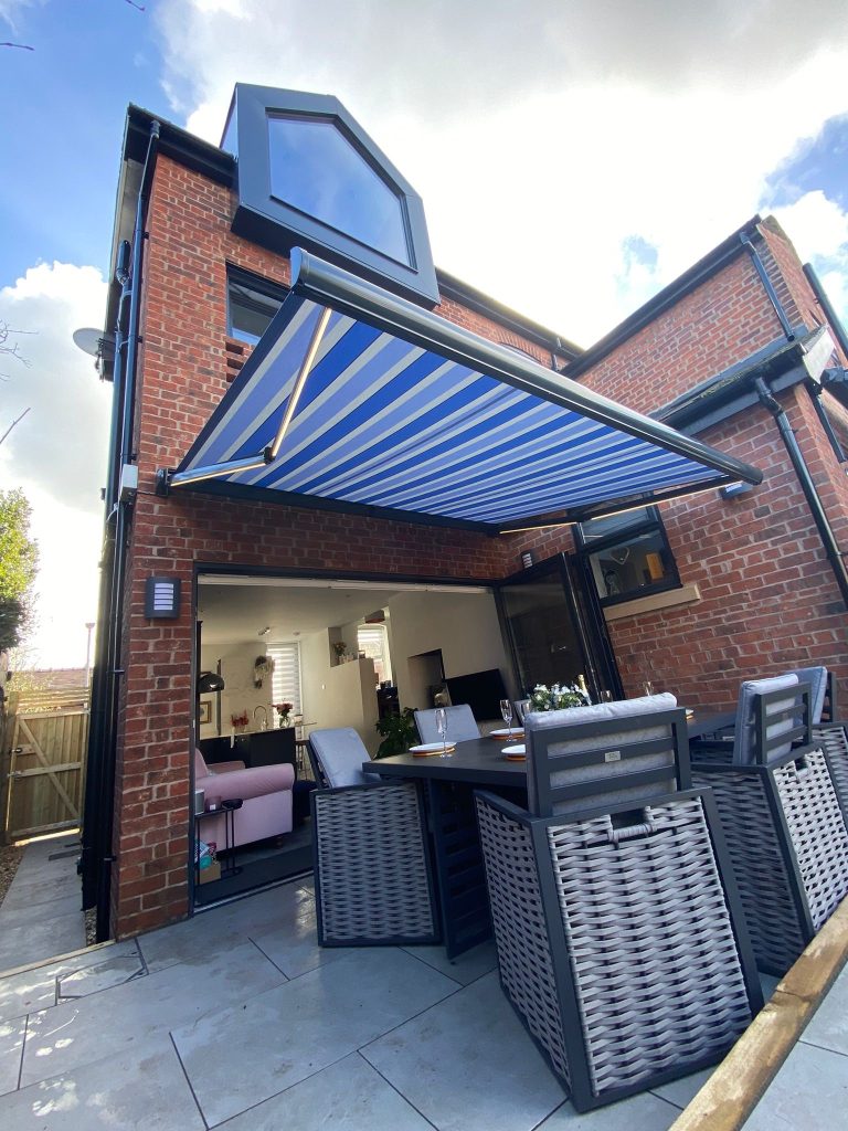 Retractable blue and white striped awning over patio dining area with bi-fold doors on brick house extension with skylight