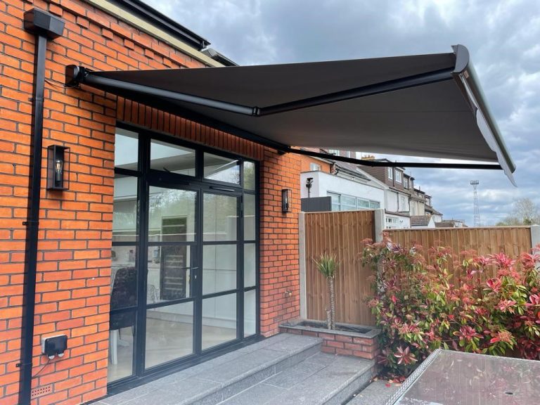 Retractable black fabric awning on brick house patio with glass doors, wooden fence, and red-leaved shrubs.