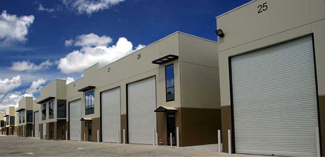 Row of numbered commercial warehouse units with white roll-up doors under a blue sky with clouds.
