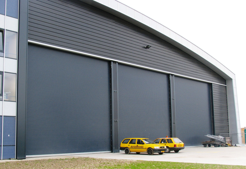 Large hangar building with dark gray corrugated roller doors and two yellow Aviodrome cars parked outside.