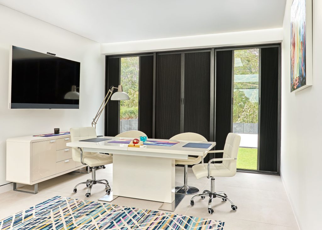 Modern home office with white desk, cream leather chairs, mounted TV, sideboard, and geometric rug near garden windows.