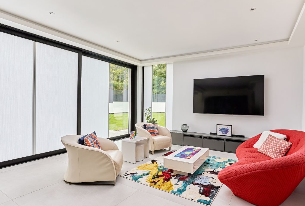 Modern living room with cream armchairs, red sofa, colorful abstract rug, wall-mounted TV, and floor-to-ceiling windows.